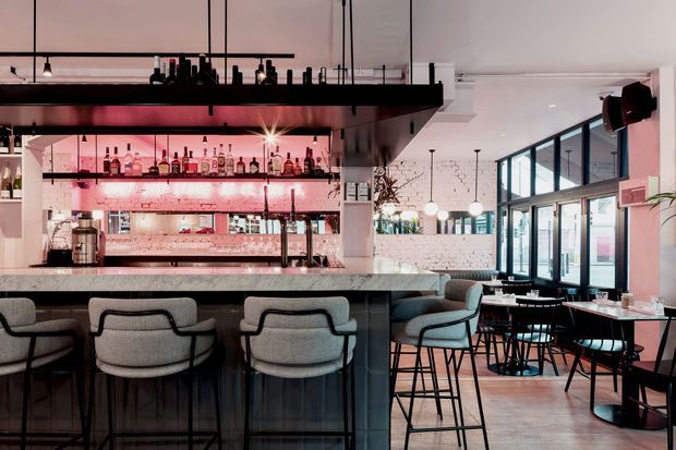 A stylish contemporary bar and restaurant interior with a prominent marble-topped bar, upholstered counter stools, illuminated liquor shelving, exposed hanging lighting, and dining seating beside large industrial-style glazed doors and windows. The palette combines pale pink lighting, white brick, dark metal, and warm timber flooring.