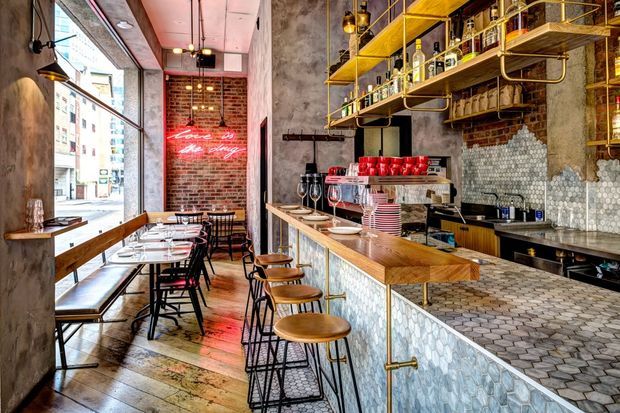 A narrow industrial-style café or bar interior with exposed brick walls, raw concrete columns, large street-facing windows, pendant lighting, timber counters and shelving, and extensive hexagonal tile finishes. The space includes a long bar with stools, café tables and chairs, coffee equipment, glassware, and a red neon sign.