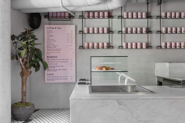 A compact contemporary café counter with a pale marble or stone-clad service island, integrated stainless-steel sink, glass pastry display, and open black metal shelving filled with pink takeaway cups. The space has grey plaster walls, exposed white ductwork, a patterned monochrome tile floor, and a large indoor potted tree. A pink menu board is mounted on the wall.