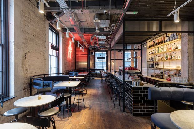 Interior view of an industrial-style bar and dining venue with exposed brick and concrete walls, an open services ceiling, timber flooring, black metal-framed glazing, communal and individual tables, bar seating, and illuminated bottle shelving. Red accent lighting adds a dramatic atmosphere.