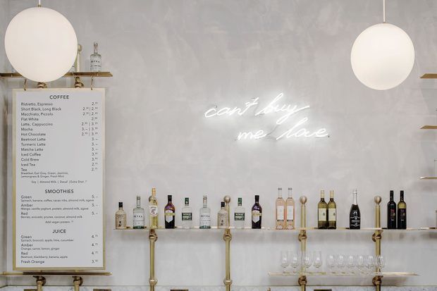 A refined beverage bar or café counter with a pale textured wall, illuminated cursive neon sign, floating glass shelves displaying bottles and glassware, brass-toned pipe supports and fittings, and large spherical pendant lights. A menu board is visible on the left.