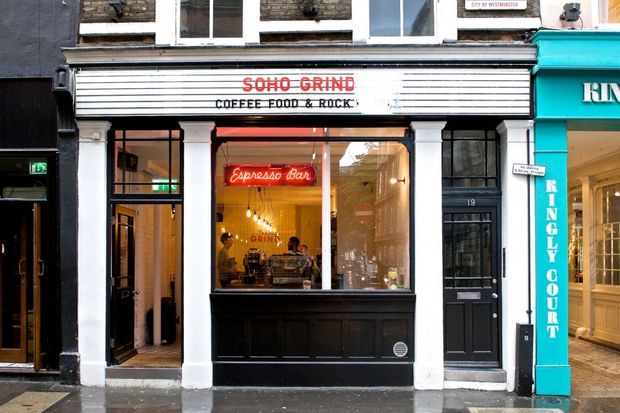 Street-level photograph of a narrow historic urban café frontage. The shop has a black-painted storefront with white pilasters, a projecting white signboard reading “SOHO GRIND COFFEE FOOD & ROCK,” large glazed windows, a neon “Espresso Bar” sign, and a black entrance door. Adjacent retail façades are visible on either side.