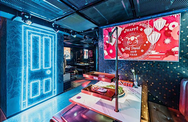 Vibrant neon-lit themed commercial dining or entertainment interior with a glowing decorative door, dark patterned wall tiles, exposed black ceiling grilles, booth seating, tables, and a large red Chinese New Year-style wall graphic. The space has a dramatic blue, red, and pink lighting scheme with an eclectic, immersive atmosphere.