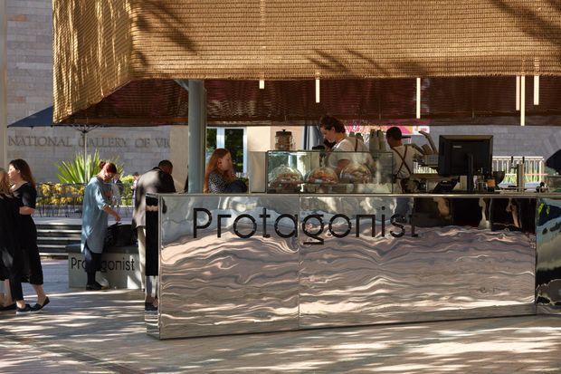 A polished outdoor or semi-outdoor café kiosk named Protagonist, positioned beside the National Gallery of Victoria. The space features a reflective undulating metal-clad service counter, glass display cases, a point-of-sale station, woven timber roller blinds overhead, and slender structural columns. Customers and staff occupy the counter area, with gallery architecture visible behind.