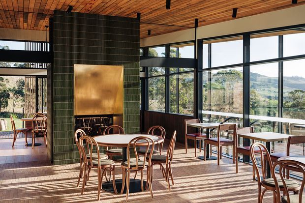 A bright, open-plan dining or café-style interior with numerous small tables and bentwood chairs. Full-height black-framed glazing overlooks a green rural landscape. The room features a warm timber slatted ceiling, timber flooring, dark green tiled feature walls, dark timber wall panelling, black structural columns, and a large brass-toned fireplace or artwork panel. Strong sunlight creates long shadows across the floor.