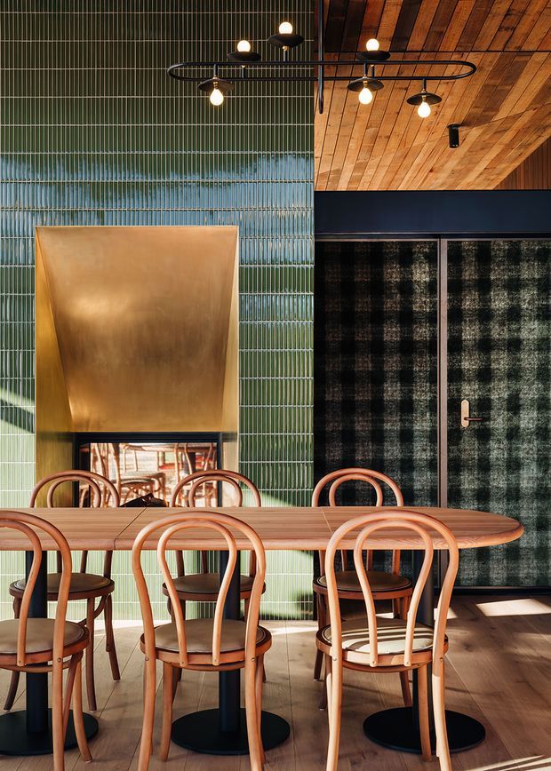 A richly styled dining room with a long timber dining table and curved timber dining chairs. The room features a dramatic green vertical-ribbed tile wall, a large brass-toned fireplace hood with firebox, dark framed patterned doors or screens, and a warm timber-lined ceiling. Sculptural black and brass pendant lighting adds a refined, moody character.