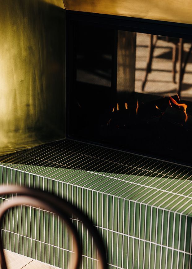 Close-up interior detail of a dark fireplace with visible flames, set above a built-in hearth clad in glossy vertically ridged green tiles with light grout. A black metal fire surround and warm wood-toned ceiling or trim create a dramatic, intimate atmosphere; a curved dark object is blurred in the foreground.