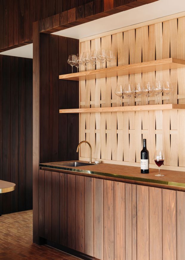 A sophisticated built-in home bar with dark timber wall panelling, a lighter timber sculptural backsplash, open glassware shelves, a brass-toned gooseneck tap, integrated sink and timber cabinetry. Wine glasses, a bottle and a glass of red wine sit on the countertop. Warm, richly textured materials create an intimate and refined atmosphere.