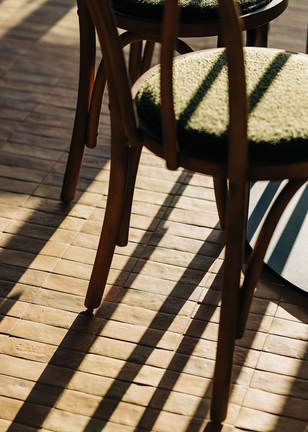 Close-up architectural detail of dark timber dining chairs with rounded legs and textured upholstered seats positioned on a warm timber deck. Strong sunlight casts long, graphic shadows across the decking, creating a warm, tactile outdoor dining atmosphere.