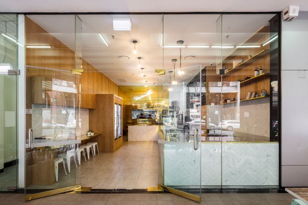 A contemporary commercial interior viewed through a fully glazed storefront. The space appears to be a café or food-service venue, with warm timber wall panelling and cabinetry, a tiled floor, glass partitions and doors, open timber shelving with displayed items, a service counter toward the rear, and bright linear and pendant lighting. The composition emphasizes the transparent shopfront and compact, modern fit-out.