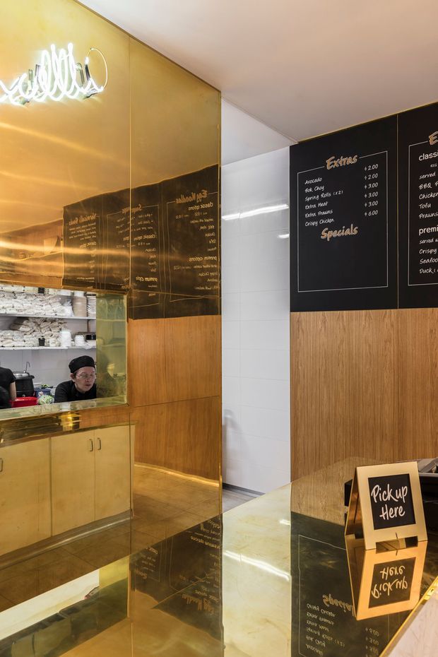 A compact commercial food-service counter or takeaway shop interior. The space features black menu boards with handwritten-style white lettering, warm brass or gold-toned reflective wall panels, timber cabinetry and wall cladding, white tiled walls, and a dark stone or polished counter. A service kitchen is visible through an opening, with a pickup sign in the foreground. The overall design is warm, refined, and slightly industrial-modern.
