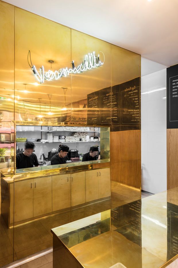 A compact commercial food-service interior with a highly reflective brass-gold wall and fixed cabinetry. A service opening reveals staff working in a stainless-steel preparation area, with shelves, appliances and stacked containers behind them. A mirrored or glossy gold-toned counter occupies the foreground, while black menu panels with white lettering and white tiled walls appear to the right. The space is brightly lit and strongly defined by reflective metallic surfaces and custom joinery.