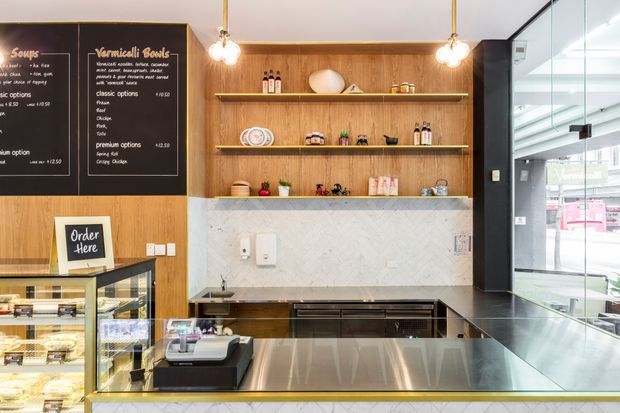 A compact contemporary commercial food-service counter, likely a café or takeaway shop. The image shows a stainless-steel preparation counter and glass display case in the foreground, dark built-in lower cabinetry, a pale veined stone splashback, and a warm timber wall with open display shelving. Chalkboard menu panels, pendant lights, service equipment, and full-height glazing are visible.