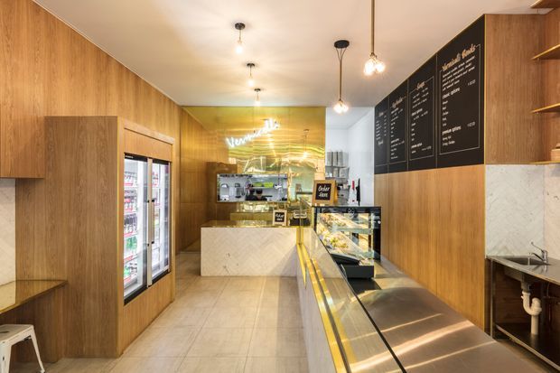 A narrow, warmly lit contemporary commercial café or dessert shop interior. The space features extensive vertical timber wall panelling and matching cabinetry, a pale tiled floor, a long glass-fronted refrigerated display counter with warm integrated lighting, a white stone service counter, black menu boards, pendant lights, and refrigerated display doors along the left wall. A small stainless-steel preparation sink is visible at the rear right.