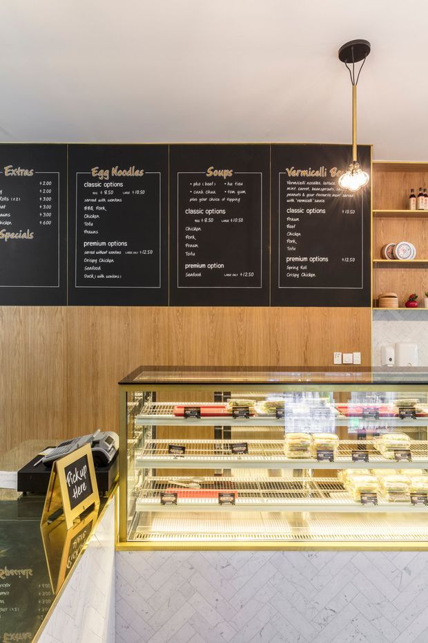 Interior view of a compact takeaway food shop or café, featuring a long illuminated refrigerated display counter, timber wall panelling, black chalkboard menu panels, open timber shelving with crockery, and a decorative pendant light. The counter front is clad in pale diagonally veined tile, creating a clean contemporary hospitality fit-out.