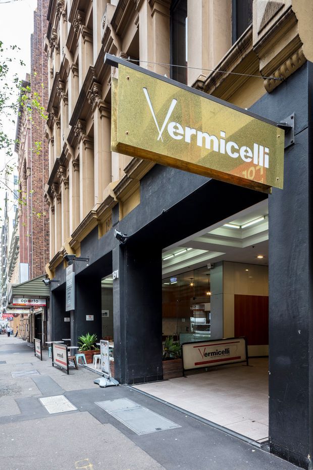 Street-level view of the Vermicelli restaurant entrance in a dense urban setting. The facade combines a dark painted storefront surround with ornate historic masonry and brick detailing above. A large projecting mustard-gold sign with white lettering identifies the venue, while the open entrance reveals a contemporary restaurant interior with a reception desk, tiled floor, timber wall panels, and layered ceiling lighting.
