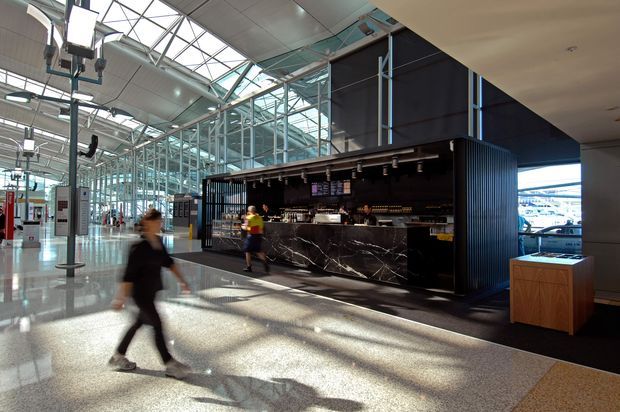 A contemporary commercial café or bar kiosk within a large, bright transport-terminal-like interior. The freestanding dark structure has ribbed black cladding, a dramatic black marble-look counter with white veining, open shelving and bar equipment. Extensive glazed roof and walls create a sunlit, airy public space, with polished terrazzo-like flooring and people moving through the foreground.