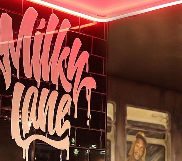 Close-up view of a dark, glossy tiled interior wall featuring a large stylised pink script mural or sign. Pink linear LED lighting runs along the ceiling perimeter, creating a dramatic red and magenta glow. A reflective or glazed surface at right shows a distorted reflection of another artwork or figure.