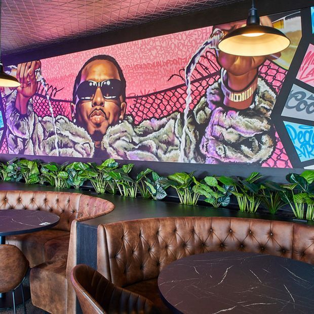 A stylish restaurant or bar interior featuring brown tufted leather banquette seating, dark round tables, a lush indoor planter strip, and a large vibrant graffiti-style mural. Black framing, pendant lights, and a patterned ceiling contribute to the dramatic urban atmosphere.