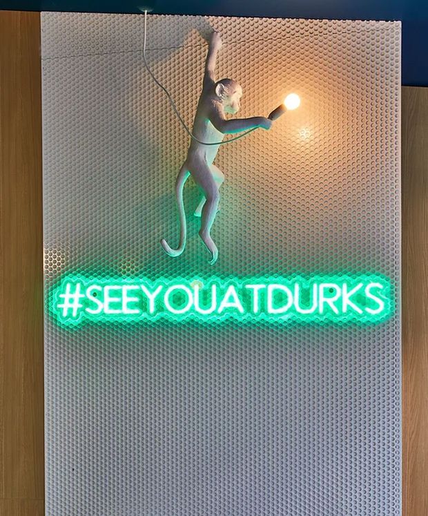 A decorative interior feature wall with a textured white panel framed by warm wood, displaying a glowing green neon hashtag sign reading “#SEEYOUATDURKS” and a monkey-shaped sculptural light fixture holding a glowing bulb. The scene has a playful, theatrical, nightlife-inspired character with dark blue surroundings.