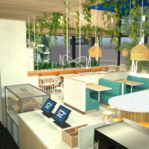 Bright contemporary commercial café or food-service interior with a pale stone service counter in the foreground, refrigerated display case, tiled counter fronts, turquoise upholstered booth seating, small dining tables, pendant lights, abundant hanging greenery, and timber ceiling and wall finishes. The space has a fresh biophilic aesthetic with white, light timber, teal, and green accents.