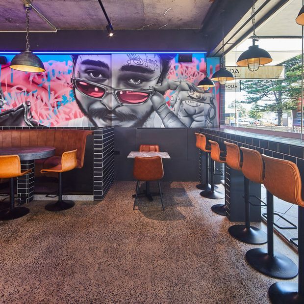 A stylish urban café or bar interior with polished terrazzo flooring, dark tiled half-height walls, timber wall panelling, black-painted ceilings, and extensive street-facing glazing. The space features leather and upholstered bar stools at a long counter, small dining tables, pendant lighting, and a large colourful graffiti-style mural with blue and pink neon lighting.