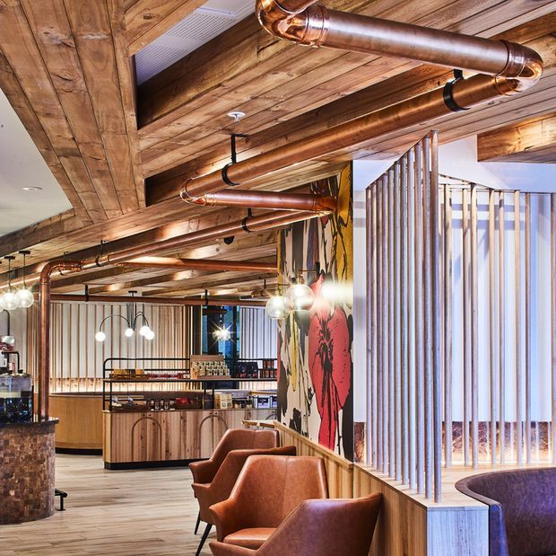 A warmly lit hospitality interior, likely a bar or restaurant, with a timber-clad ceiling, exposed copper pipework, vertical timber screening, pendant lighting, timber service cabinetry, leather lounge chairs, and a bold floral wall mural. The space has an industrial yet refined character with strong natural wood and copper detailing.