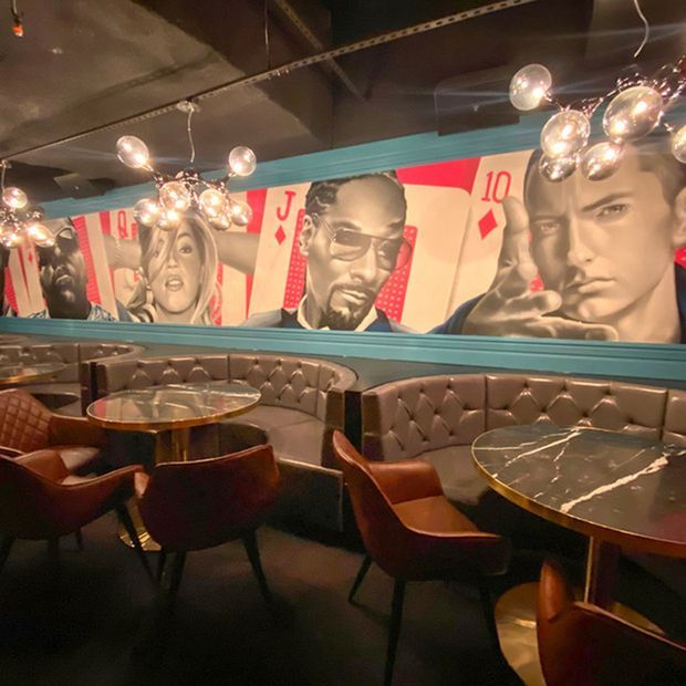A moody, richly decorated hospitality interior with curved, tufted banquette seating, brown leather armchairs, round marble-topped tables, and clustered globe pendant lights. A large pop-art mural featuring playing-card imagery and portrait figures spans the wall, framed by turquoise trim beneath a dark exposed ceiling.
