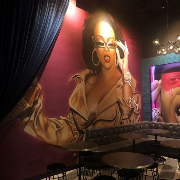 A dramatic, dimly lit entertainment or hospitality interior featuring a large colourful mural of a woman in sunglasses, deep burgundy walls, black drapery, tufted banquette seating, small round tables, stools, and a glowing wall-mounted screen or artwork. A sculptural cluster pendant light adds a decorative focal point above the seating area.