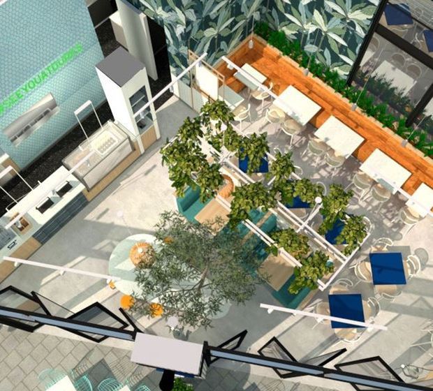 Aerial interior view of a bright, contemporary commercial atrium or hospitality space. The open-plan area contains multiple white tables and chairs, planted outdoor-style seating zones, raised garden beds with trees and shrubs, pale tiled flooring, timber wall lining, and extensive glass and angular black-framed glazing. A turquoise tiled feature wall and service counter are visible at the left.