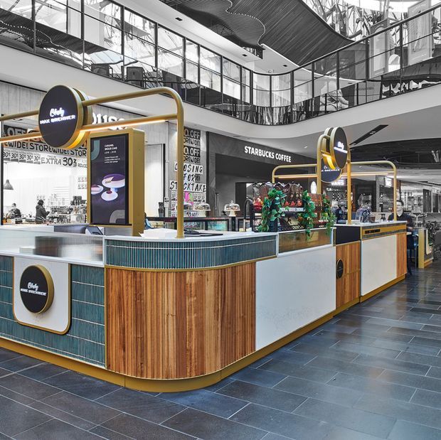 A contemporary shopping-centre food and beverage kiosk, with a Starbucks Coffee counter as the central subject. The kiosk features curved counters, vertical timber cladding, dark teal ribbed tile panels, white solid-surface sections, and brass-coloured tubular frames and trims. Large glazed mall interiors, upper-level walkways, and retail signage are visible in the background. The space is brightly lit with a polished, high-traffic commercial character.