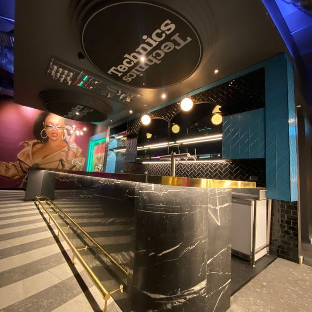 A dramatic, dark-toned commercial bar or hospitality interior with a curved black marble-clad counter, brass edging and handrails, teal geometric wall tiles, black herringbone tile backsplash, glowing pendant lights, mirrored shelving, and a large mural. A circular ceiling feature displays the venue branding.