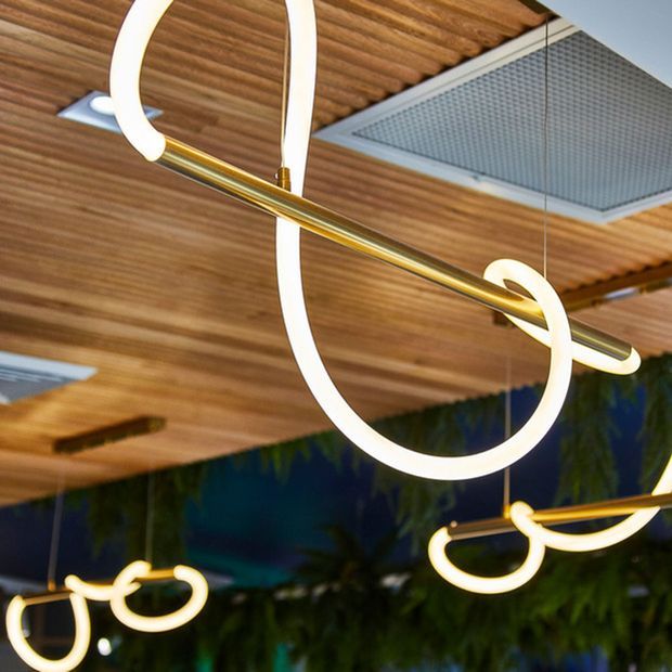 Close-up architectural detail of sculptural contemporary pendant lighting with looping illuminated forms and brass-toned horizontal rods, suspended beneath a warm timber slatted ceiling. Recessed linear lighting and a dark reflective or glazed background are also visible, creating a refined modern atmosphere.