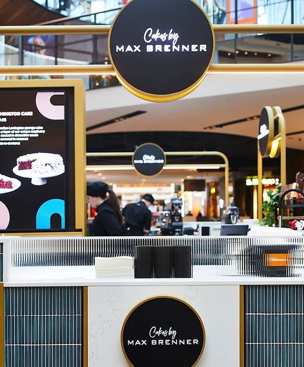 A Max Brenner chocolate café kiosk inside a modern shopping centre, with prominent circular black-and-gold branding, a digital dessert menu, a white marble-look service counter, and dark vertical tile detailing. Retail activity and mall balconies are visible in the background.