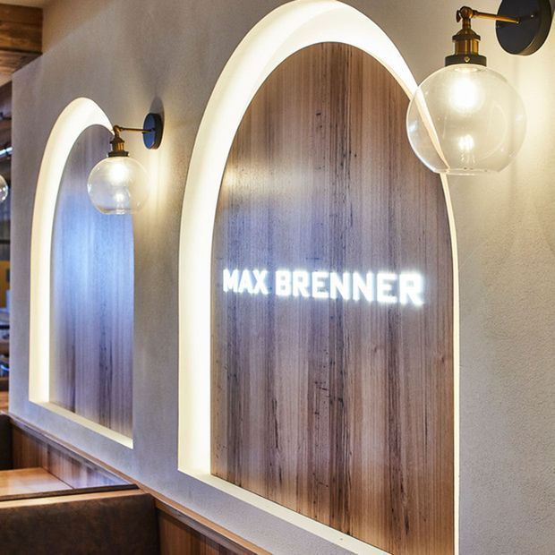 Close-up interior architectural detail of a Max Brenner commercial chocolate restaurant, featuring arched illuminated wall niches with vertical timber-panelled backings, warm globe wall sconces, textured pale plaster walls, and built-in booth seating.