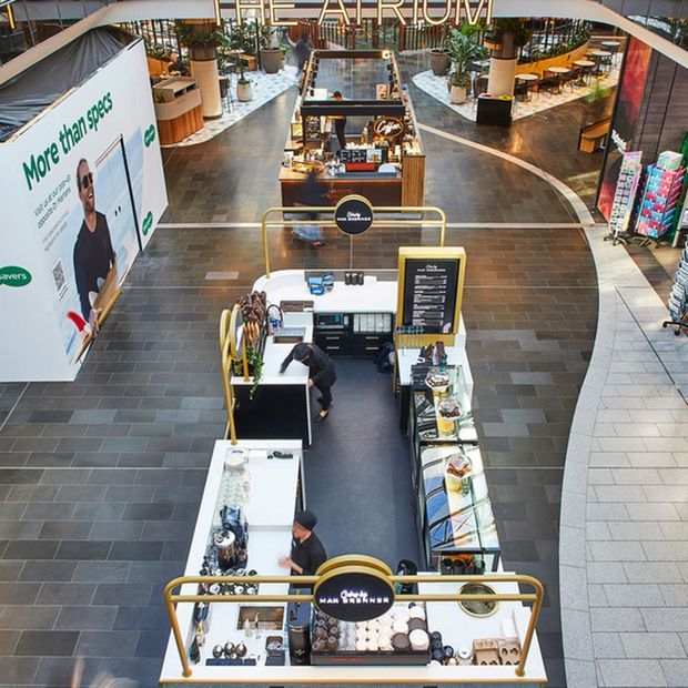 Elevated view of a contemporary indoor shopping centre atrium with multiple compact café and retail kiosks arranged along a broad tiled concourse. The space features dark grey and light grey tiled flooring, curved circulation paths, planted seating areas, timber and white kiosk joinery, and prominent overhead signage and advertising.