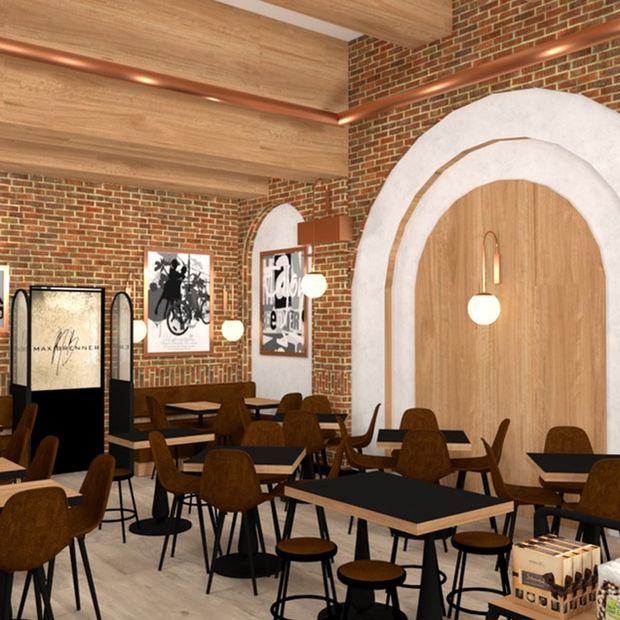 A warm, brick-lined café or restaurant interior with numerous dark wood tables and brown upholstered chairs. The space features a dramatic arched timber-clad wall niche or doorway framed in white plaster, exposed red brick walls, timber ceiling beams and panels, copper-toned linear detailing, pendant globe lights, and framed artwork.