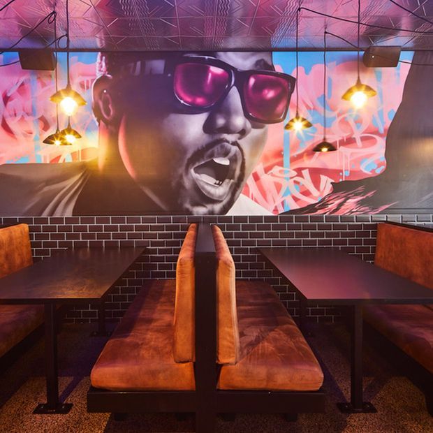 A vibrant urban-style restaurant or bar interior with dark brick walls, booth seating upholstered in warm cognac leather, dark timber tables, pendant lights, and a large colourful pop-art mural featuring a musician wearing sunglasses. The ceiling has an expressive geometric patterned finish, creating a bold, energetic atmosphere.