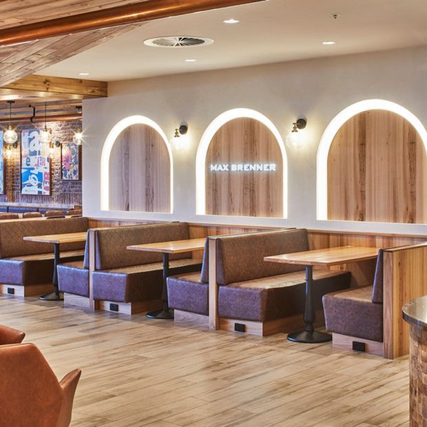 A warm, polished restaurant interior with rows of upholstered booth seating, timber tables, light timber flooring, and a feature wall with three illuminated arched timber panels. Exposed timber ceiling elements, pendant lights, wall sconces, and framed artwork create a welcoming hospitality setting.