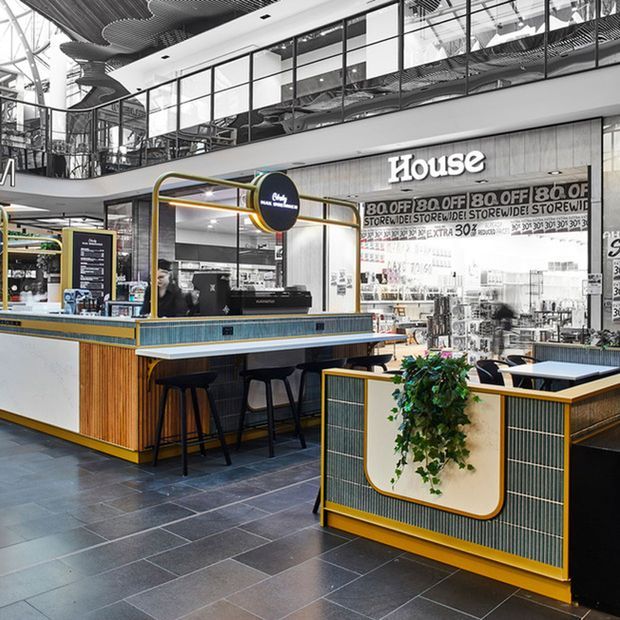 A contemporary indoor commercial food-and-beverage kiosk or café within a large retail or shopping-centre environment. The space features teal and pale blue vertically ribbed fixed joinery, warm timber slats, mustard-gold metal framing and trims, dark bar stools, and dark large-format tiled flooring. A curved counter enclosure with integrated planting sits in the foreground, while open upper-level glazing, exposed services, and a prominent House retail frontage are visible behind.