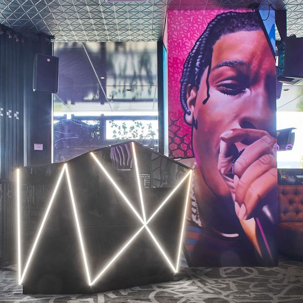 A dramatic commercial interior featuring a dark reception or DJ-style counter with bright geometric linear LED lighting, a large colourful mural of a musician, a patterned geometric ceiling, dark wall finishes, and patterned carpet. Seating and glazing are visible in the background.