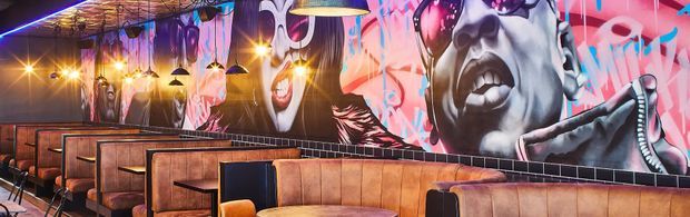 A vibrant commercial dining or bar interior with rows of upholstered booth seating, dark tabletops, hanging pendant lights, exposed ceiling services, and large colourful graffiti-style murals covering the walls. The space has a bold urban atmosphere with warm ambient lighting and industrial-inspired finishes.