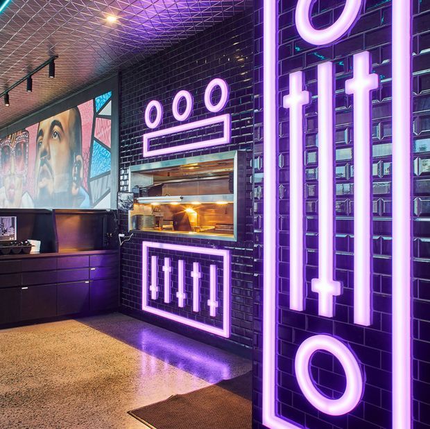 A dramatic, neon-lit commercial interior with glossy black brick walls, large purple and white illuminated graphic symbols, a built-in service or display opening, dark cabinetry, artwork, and a reflective polished floor. The ceiling has a geometric patterned finish with track lighting, creating a bold entertainment or hospitality atmosphere.