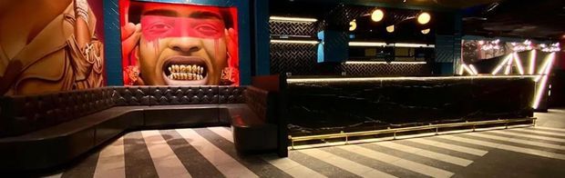 A dark, dramatic commercial interior resembling a nightclub or bar, with a long black tufted banquette wrapping around the room, a large colorful mural, glossy black surfaces, bright ceiling spotlights, and bold black-and-white striped flooring.