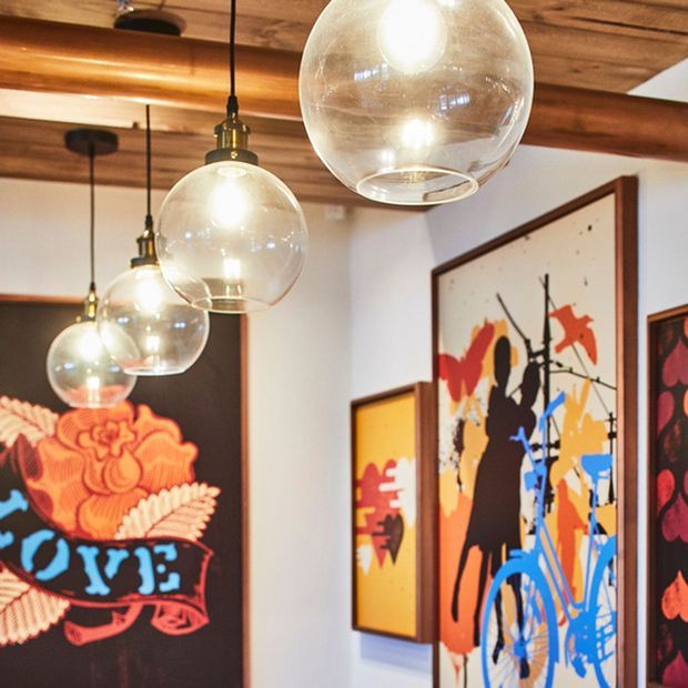 Close-up interior view of a warmly lit room featuring a timber-lined ceiling with exposed beams, a row of suspended clear glass globe pendant lights, and colourful framed abstract artwork arranged along white walls. The space has an eclectic, gallery-like character with strong graphic artwork and warm wood tones.