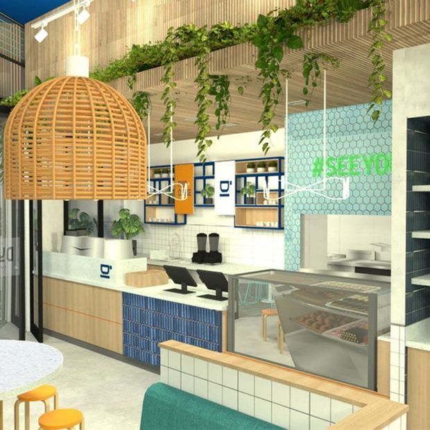 A bright contemporary commercial café or food-service interior with a visible service counter, pastry display case, shelving, tiled surfaces, and branded green lettering. The space features a large woven timber pendant light, suspended greenery, blue and white cabinetry, pale timber wall cladding, and a patterned hexagonal tile feature wall.
