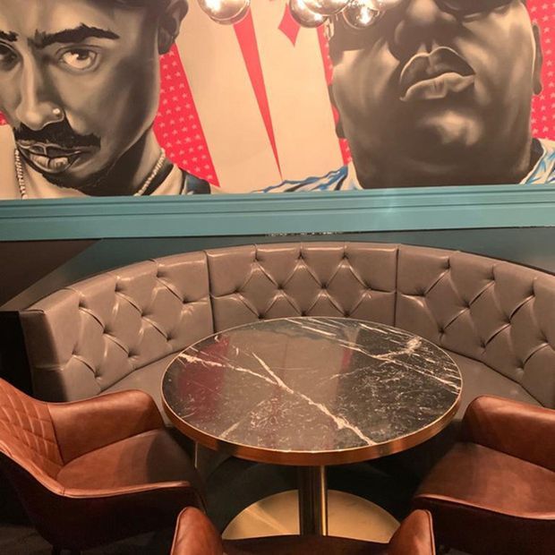 A stylish restaurant or lounge booth with a curved, tufted brown leather banquette surrounding a round dark marble-topped table, paired with rust-colored upholstered armchairs. A teal wall trim and large bold pop-art portraits create a dramatic, retro-inspired atmosphere, with pendant globe lighting overhead.