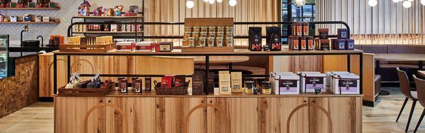 A warm, wood-lined commercial interior resembling a café or specialty food and beverage shop. The image shows a long timber service and display counter with curved panel detailing, shelving stocked with packaged goods, countertop equipment, pendant globe lighting, and a seating area at the right. Vertical timber slats and dark-framed glazing add texture and structure.