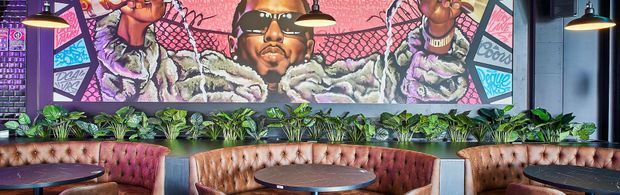 A stylish commercial lounge or hospitality interior featuring a long tufted leather banquette in warm brown and burgundy tones, round dark tables, pendant lights, lush planter greenery, and a large vibrant graffiti-style mural across the rear wall. The space has a bold, urban atmosphere with dark architectural finishes and colourful artwork.