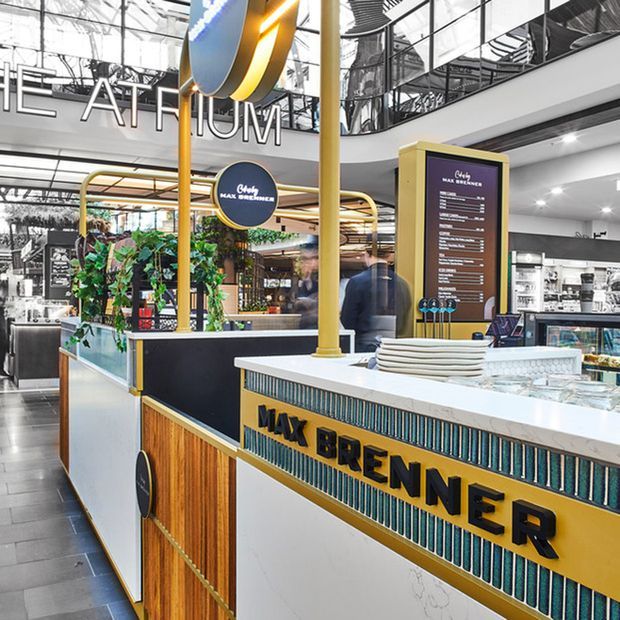Interior view of a Max Brenner chocolate café kiosk in a shopping centre, featuring a prominent white service counter with bold yellow and black branding, teal vertical tile detailing, timber panelling, menu boards, display shelving, and decorative planting. The surrounding multi-level mall structure and glazed upper-level balustrades are visible in the background.